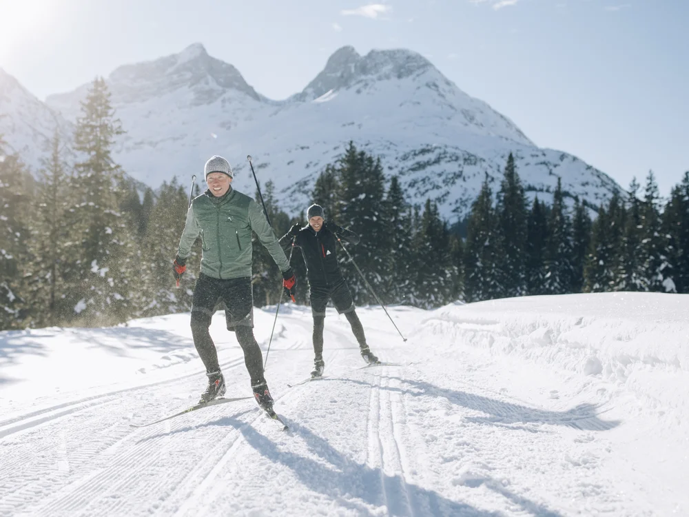 Ihre Auszeit in Lech am Arlberg Zwei Langläufer auf einer verschneiten Loipe vor bewaldeten Bergen