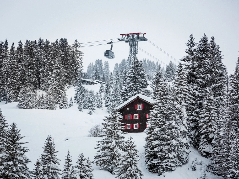 Ihre Auszeit in Lech am Arlberg Verschneite Tannen umgeben ein Chalet mit roten Fensterläden und eine Gondelbahn