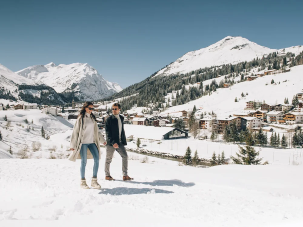 Ihre Auszeit in Lech am Arlberg Paar spaziert durch eine verschneite Berglandschaft mit einem Dorf im Hintergrund