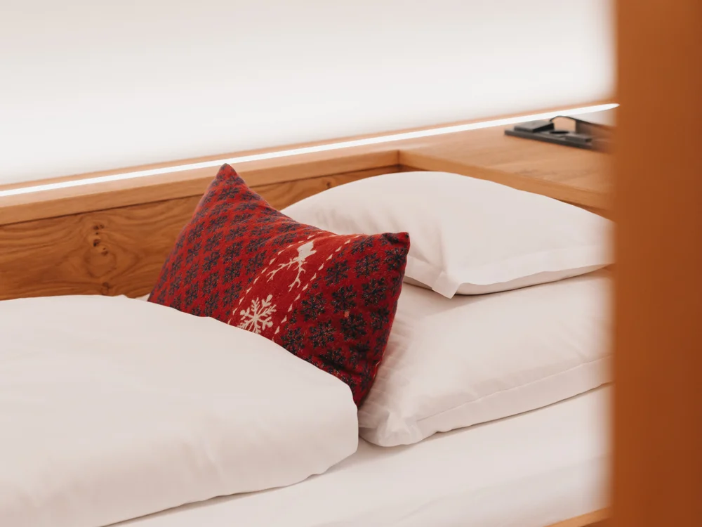 Ihre Auszeit in Lech am Arlberg Bett aus Holz mit weißer Bettwäsche und rotem gemustertem Kissen