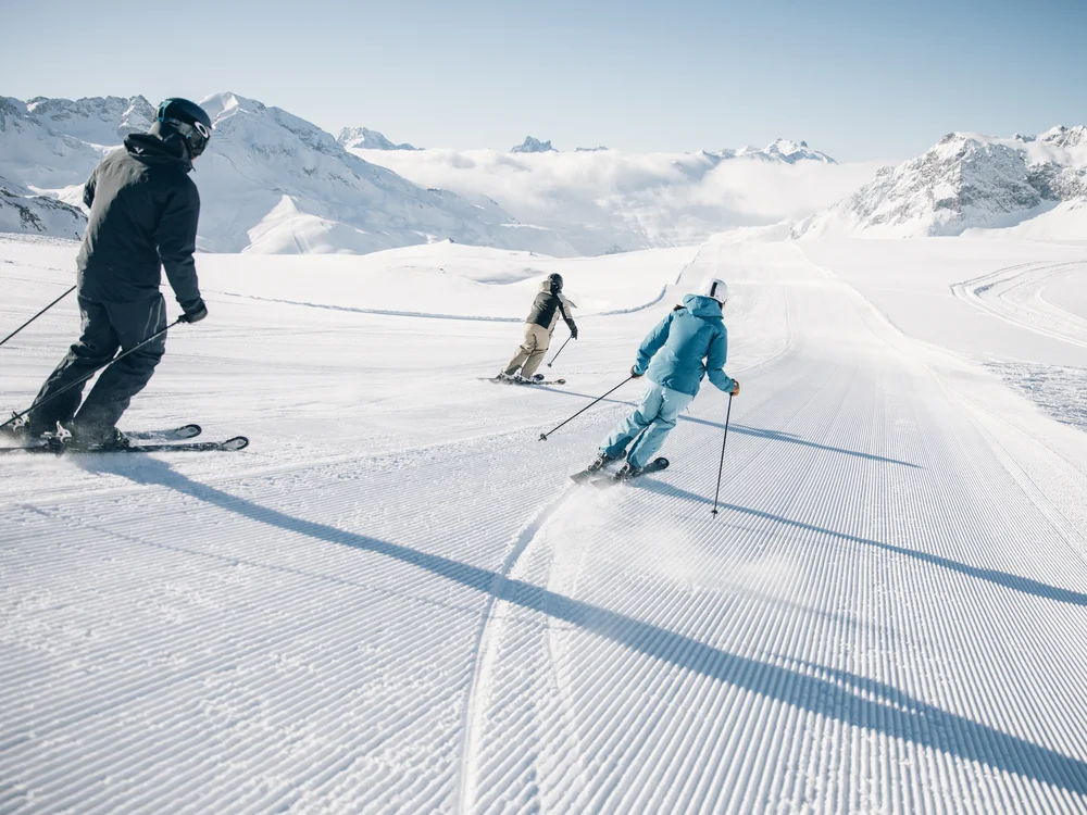 Ihre Auszeit in Lech am Arlberg Drei Skifahrer fahren auf einer präparierten Piste vor verschneiten Bergen