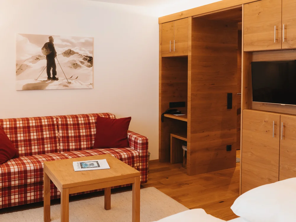 Ihre Auszeit in Lech am Arlberg Wohnbereich mit kariertem Sofa, Holzmöbeln, Couchtisch und sichtbarem Bett