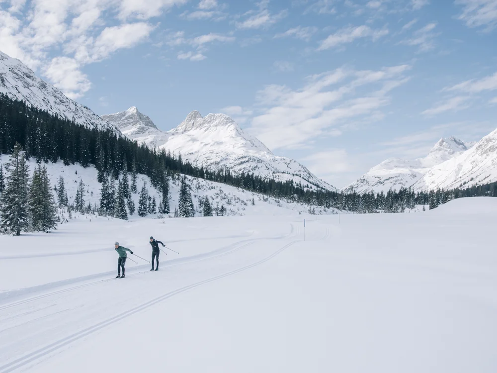 Ihre Auszeit in Lech am Arlberg Zwei Langläufer gleiten durch ein verschneites Tal vor schneebedeckten Bergen