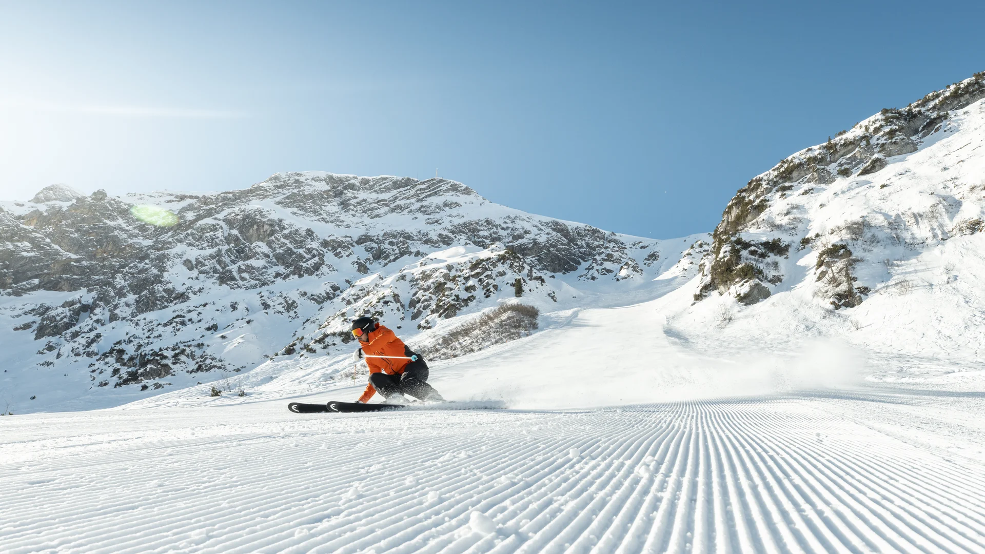 Skifahrer in orangefarbener Jacke fährt eine präparierte Piste vor schneebedeckten Bergen hinab