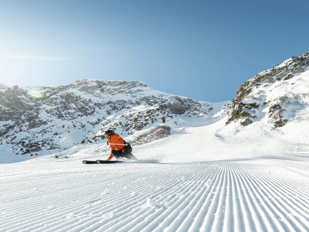 Ihre Auszeit in Lech am Arlberg Skifahrer in orangefarbener Jacke fährt eine präparierte Piste vor schneebedeckten Bergen hinab