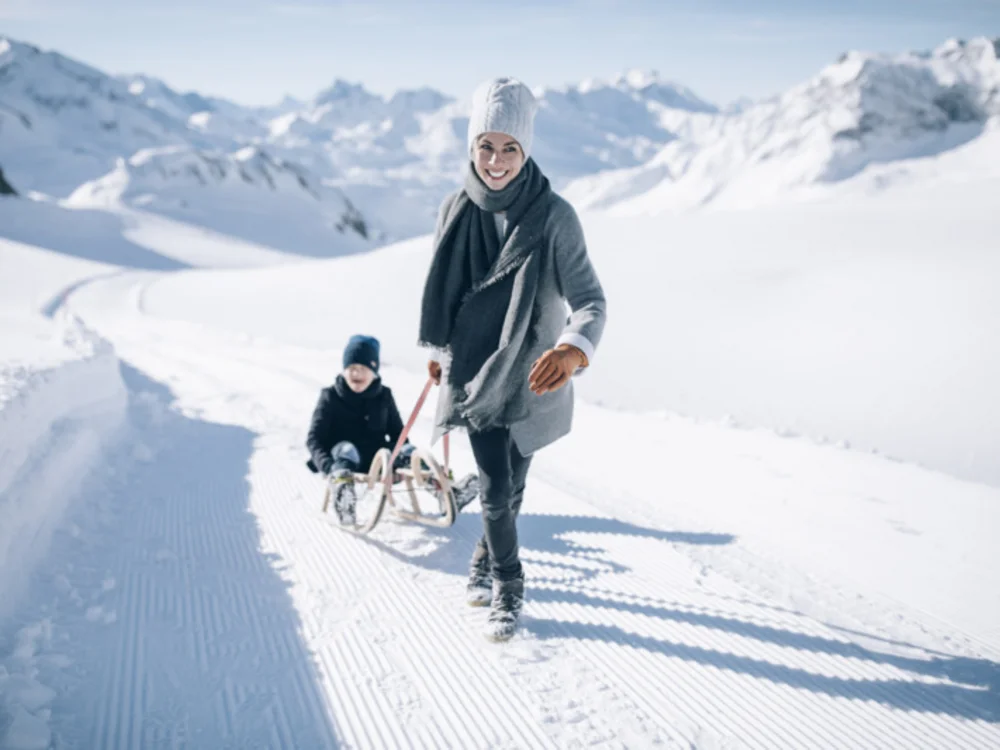 Ihre Auszeit in Lech am Arlberg Frau zieht ein Kind auf einem Schlitten über einen verschneiten Weg in den Bergen