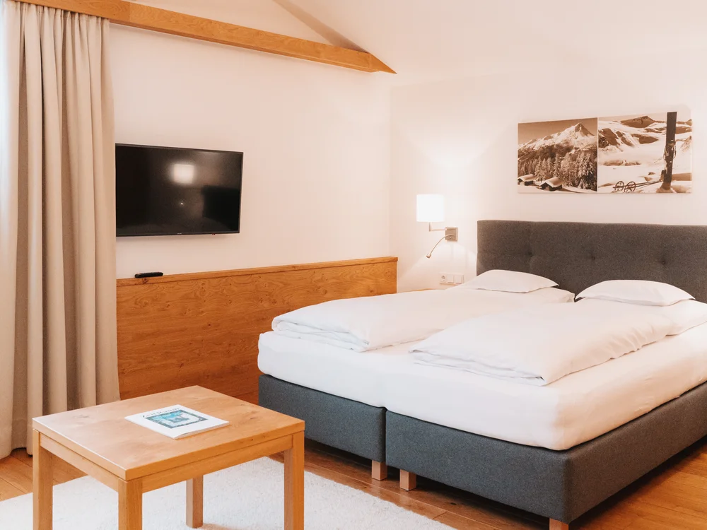 Ihre Auszeit in Lech am Arlberg Helles Schlafzimmer mit Doppelbett, Holztisch, Fernseher und Bergbild an der Wand