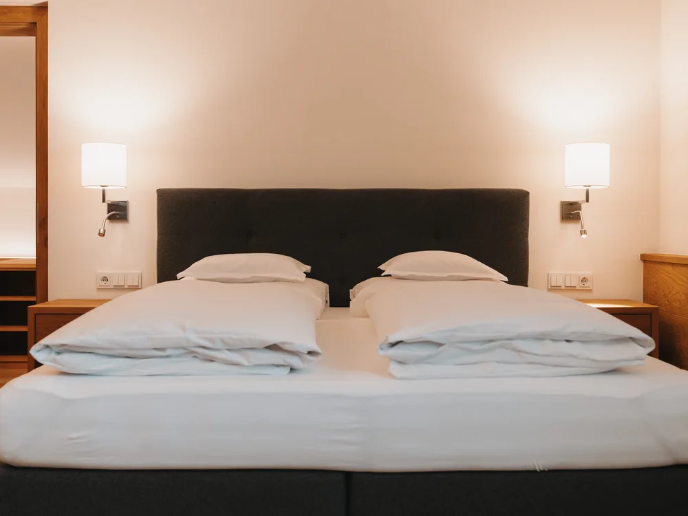 Ihre Auszeit in Lech am Arlberg Hotelbett mit zwei Einzeldecken, schwarzem Kopfteil und zwei Wandleuchten