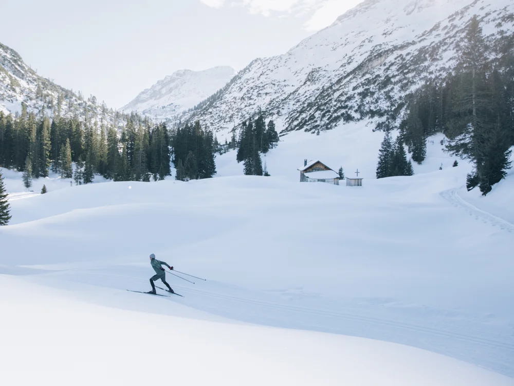 Ihre Auszeit in Lech am Arlberg Langläufer auf einer verschneiten Ebene vor bewaldeten Bergen und einer kleinen Hütte
